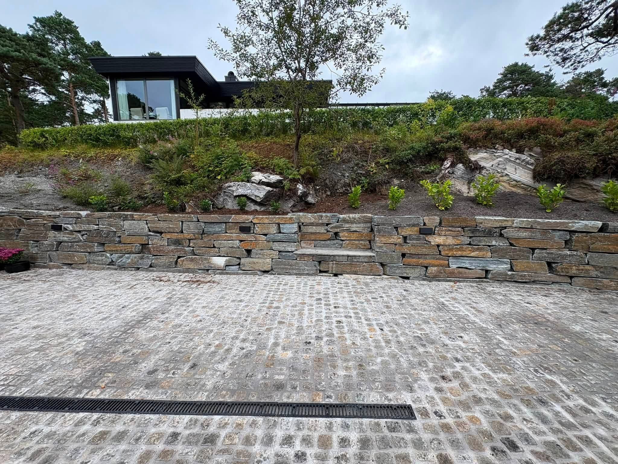 Natursteinsmur med beplantning over dekket av brukt smågatestein